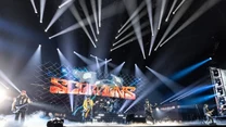 Zespół muzyczny występuje na scenie oświetlonej efektownymi światłami, w tle duży ekran z nazwą Scorpions, na scenie widoczne instrumenty, muzycy oraz dym sceniczny podkreślający koncertowy nastrój.