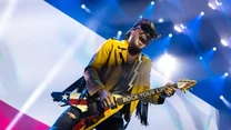 Mężczyzna w żółtej kurtce i okularach przeciwsłonecznych gra na gitarze elektrycznej o wzorze amerykańskiej flagi podczas koncertu, wokół niego jasne światła sceniczne.