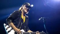 Muzyk grający na elektrycznej gitarze, ubrany w czarną koszulę z żółtym akcentem oraz czapkę z daszkiem, dynamiczne światło sceniczne oświetla postać oraz gitarę zdobioną biało-czarnym wzorem, w tle mikrofon.
