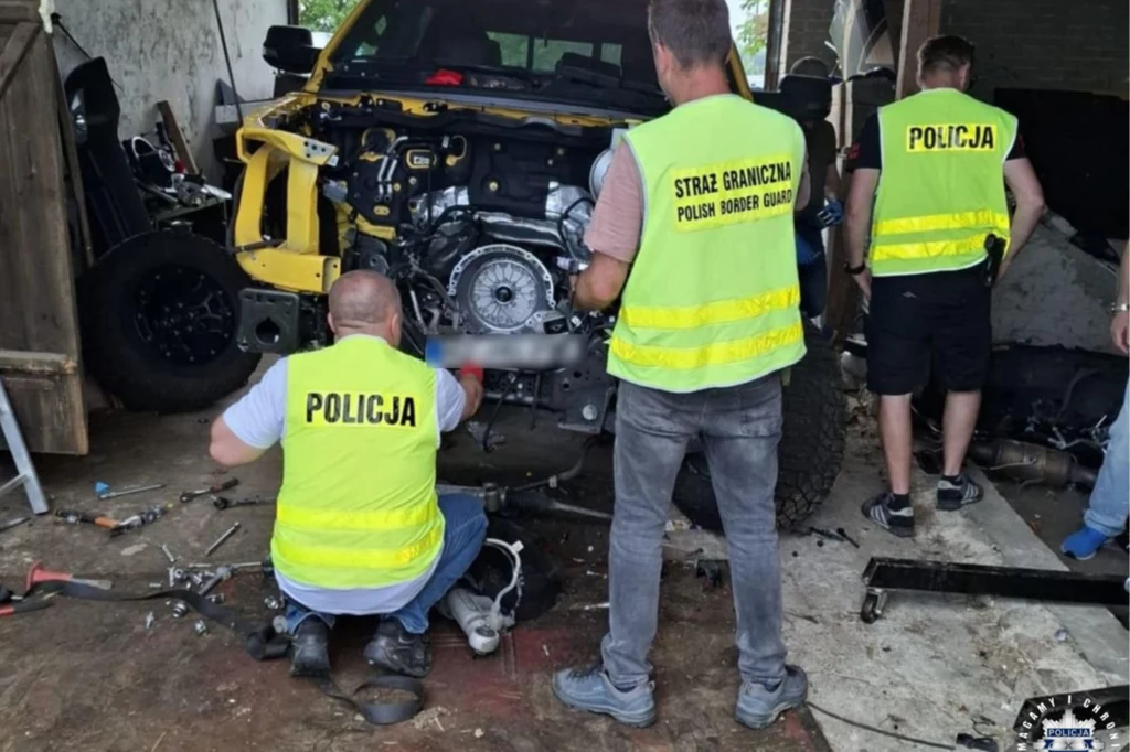 Policjanci zabezpieczyli ponad 40 pojazdów różnych marek. Funkcjonariusze Policji i Straży Granicznej w żółtych kamizelkach z napisami kontrolują oraz rozbierają samochód terenowy na terenie warsztatu, wokół widoczne części samochodowe oraz narzędzia.