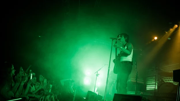 Wokalista w białej koszulce i z gitarą występuje na scenie oświetlonej zielonym światłem, publiczność stoi blisko barierek i nagrywa występ telefonami.