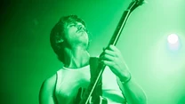 Mężczyzna grający na gitarze elektrycznej na scenie, oświetlony zielonym światłem, dynamiczna poza, skoncentrowany wyraz twarzy, tło rozmyte przez intensywne oświetlenie sceniczne.