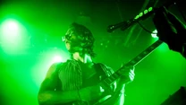 Muzyk grający na gitarze elektrycznej podczas koncertu, silnie oświetlony zielonym światłem sceny, dynamiczna poza, widoczny mikrofon oraz fragment innego sprzętu scenicznego.