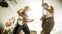 Trzech muzyków grających na gitarach elektrycznych na jasno oświetlonej scenie, w tle widoczne instrumenty i sprzęt muzyczny.