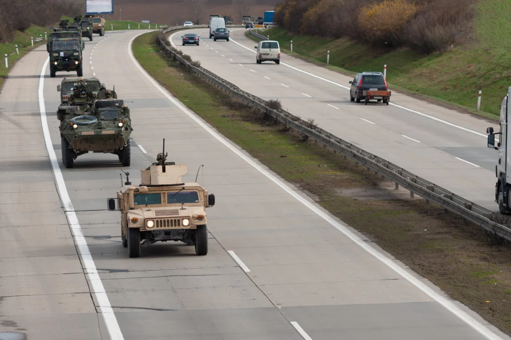 Konwój wojskowych pojazdów opancerzonych poruszający się autostradą w towarzystwie cywilnych samochodów, widoczny podział dróg oddzielający oba kierunki ruchu, przydrożne drzewa oraz prosta linia żywej zieleni po jednej stronie drogi.