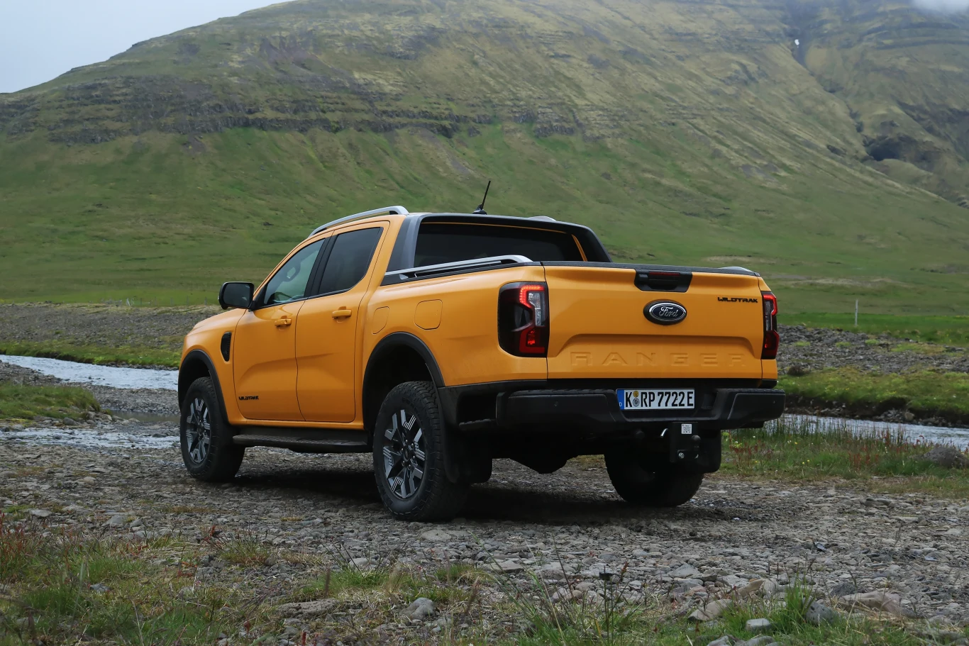 Pomarańczowy pickup Ford Ranger stoi na kamienistej drodze tuż obok małego, płytkiego strumienia, z tyłu widać zielone, łagodne wzgórza porośnięte trawą.
