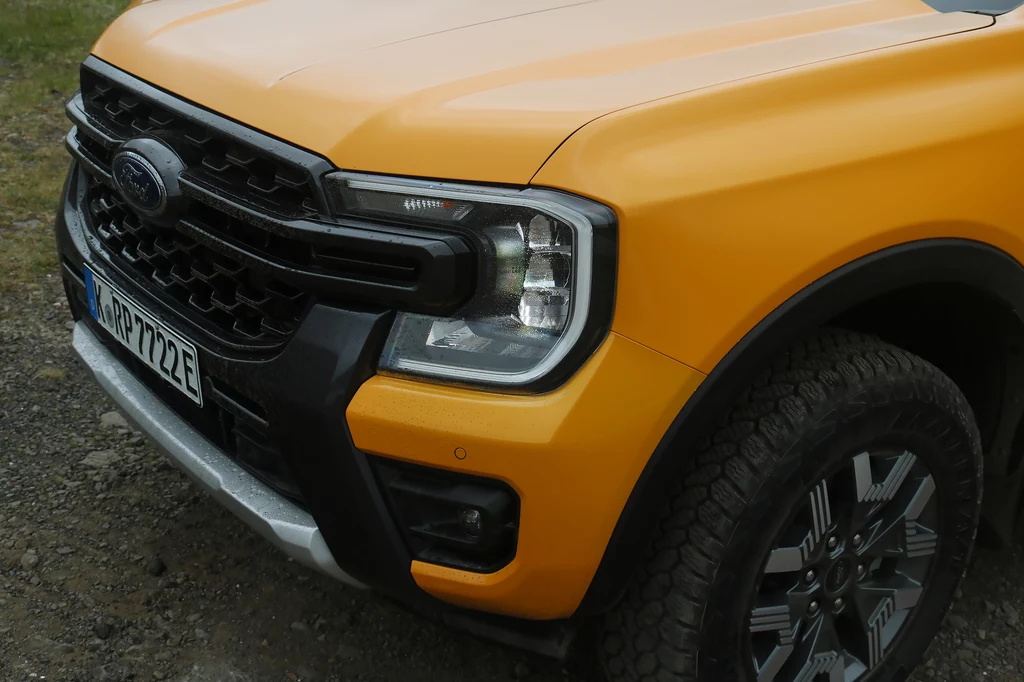 Układ napędowy potrafi automatycznie rozdzielać moment obrotowy z obu na wszystkie cztery koła. Fragment żółtego samochodu terenowego marki Ford, widoczna maska, grill, reflektor, zderzak oraz przednie koło z charakterystyczną felgą, pojazd stoi na kamienistym podłożu.