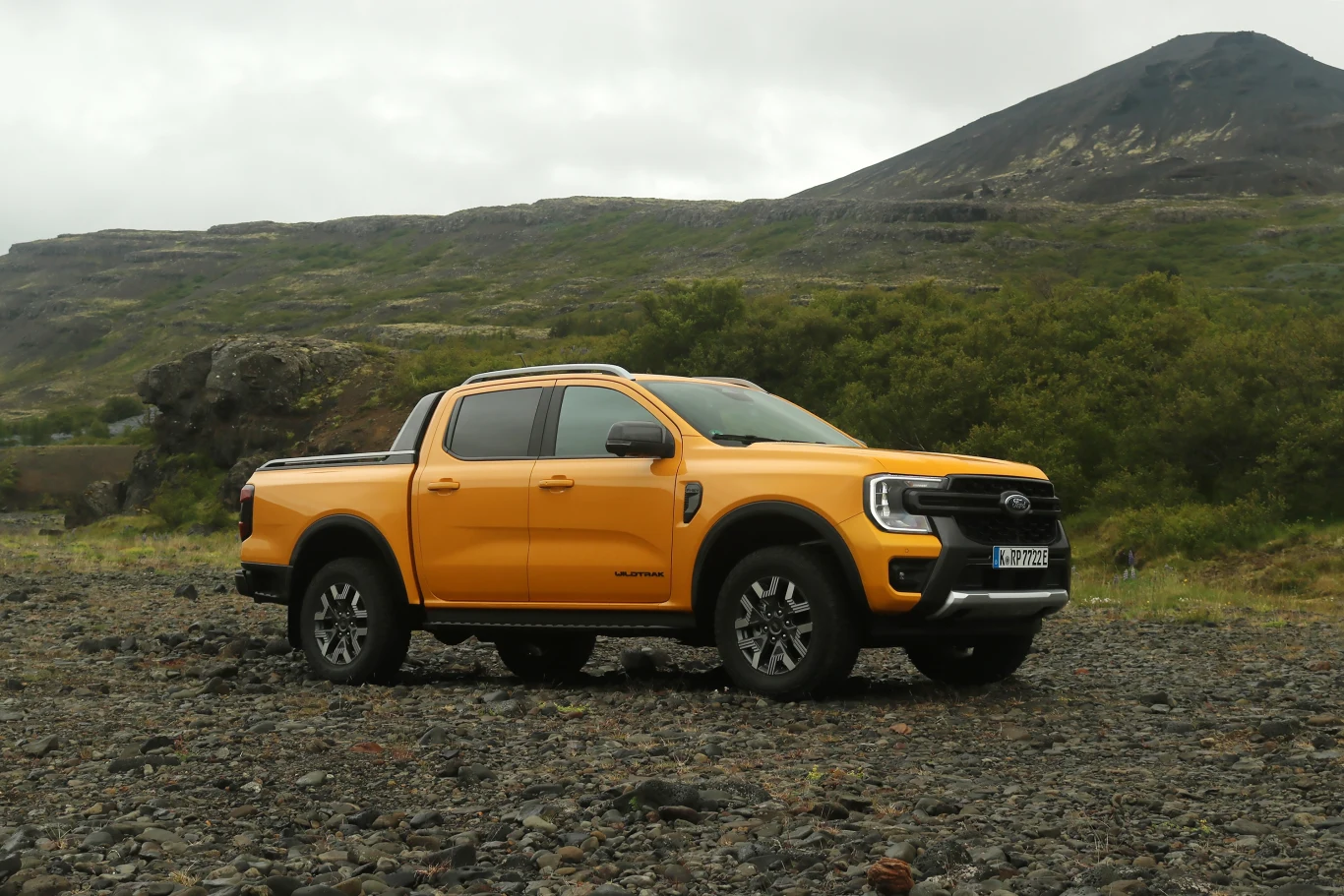 Pomarańczowy pickup z napędem na cztery koła stoi na kamienistym terenie, w tle widoczne są zielone krzewy i wzgórza pokryte trawą oraz szczyt górski, niebo jest zachmurzone.