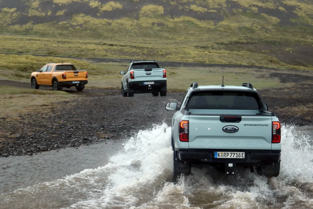Ford Ranger Plug-in przejedzie przez kamienie, błoto i rzeki. Trzy pickupy Forda pokonują kamienistą i zalaną drogę w górzystym, zielonym krajobrazie, podczas jazdy terenowej, widoczne są rozpryski wody oraz surowa przyroda.