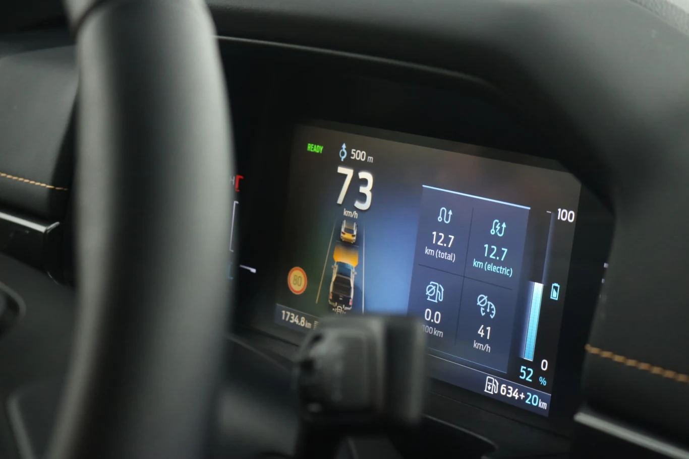 Wyświetlacz cyfrowy samochodu z widoczną prędkością 73 km/h, wskaźnikiem zużycia energii, stanem baterii i przebiegiem, częściowo widoczna kierownica po lewej stronie.