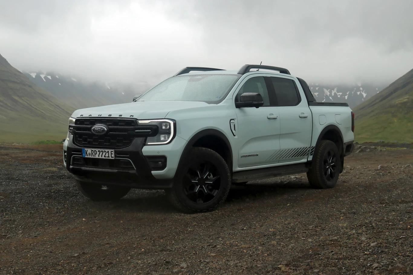 Jasnoszary pickup marki Ford w terenie górskim, otoczony mgłą i niskimi chmurami, stoi na nierównym, żwirowym podłożu, w tle widoczne strome zbocza pokryte resztkami śniegu.