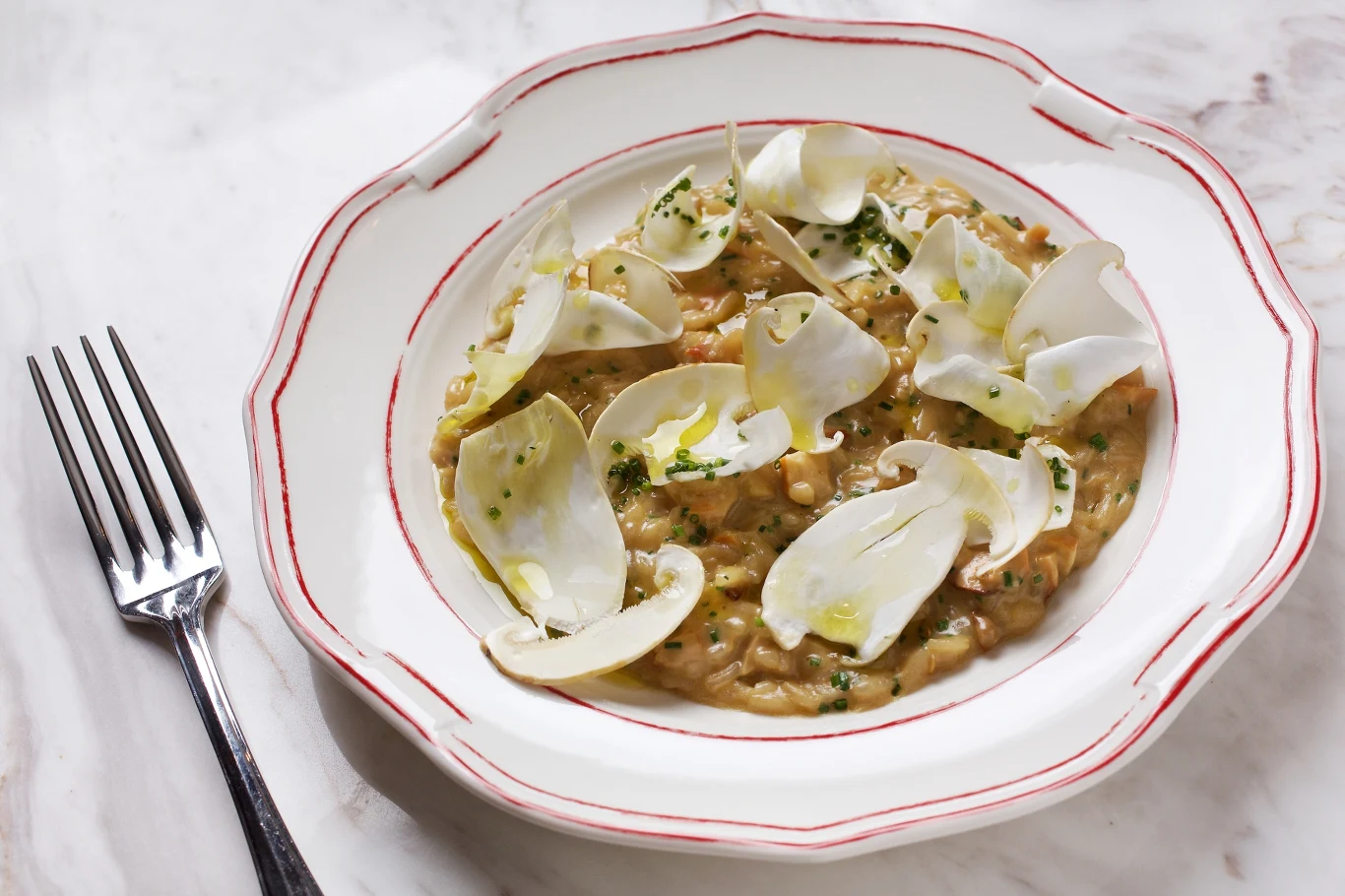 Risotto Acquerello Carnaroli z grzybami: żółciakiem siarkowym i matsutake, pietruszką i parmezanem, sfotografowane w restauracji Officina w Waszyngtonie Kremowe risotto z cienko pokrojonymi białymi grzybami ułożonymi na wierzchu, polane oliwą i posypane świeżymi ziołami, podane na eleganckim, ozdobnym talerzu z czerwoną linią na brzegu.