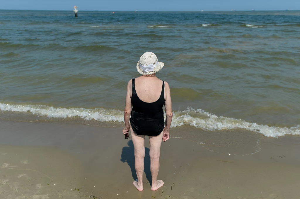 Seniorzy powinni pamiętać, by na plaży ostrożnie dozować sobie wysiłek fizyczny Starsza kobieta w czarnym stroju kąpielowym i białym kapeluszu stoi na brzegu morza, patrząc na spokojne fale przy linii brzegowej.