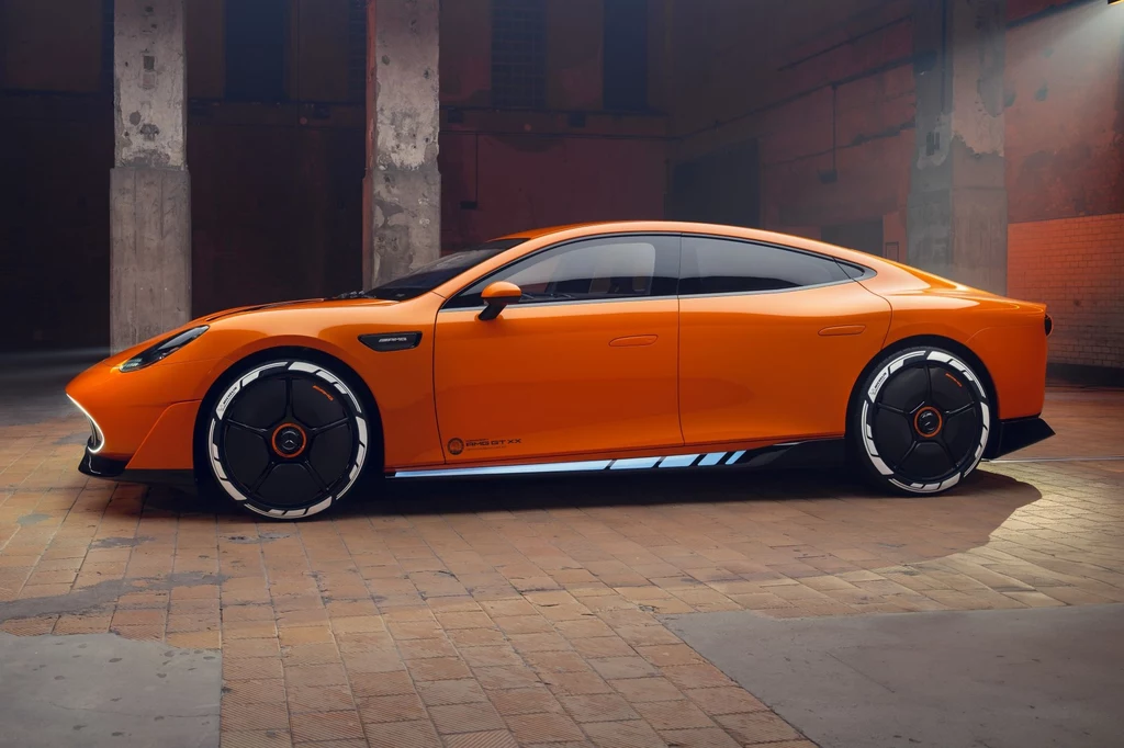 Współczynnik oporu powietrza (Cd) w Concept AMG GT XX wynosi 0,198. Pomarańczowy, sportowy samochód elektryczny o nowoczesnym designie stoi zaparkowany w pustym, industrialnym wnętrzu o surowych, betonowych ścianach i podłodze z cegły. Charakterystyczne felgi i aerodynamiczna sylwetka podkreślają futurystyczny charakte...