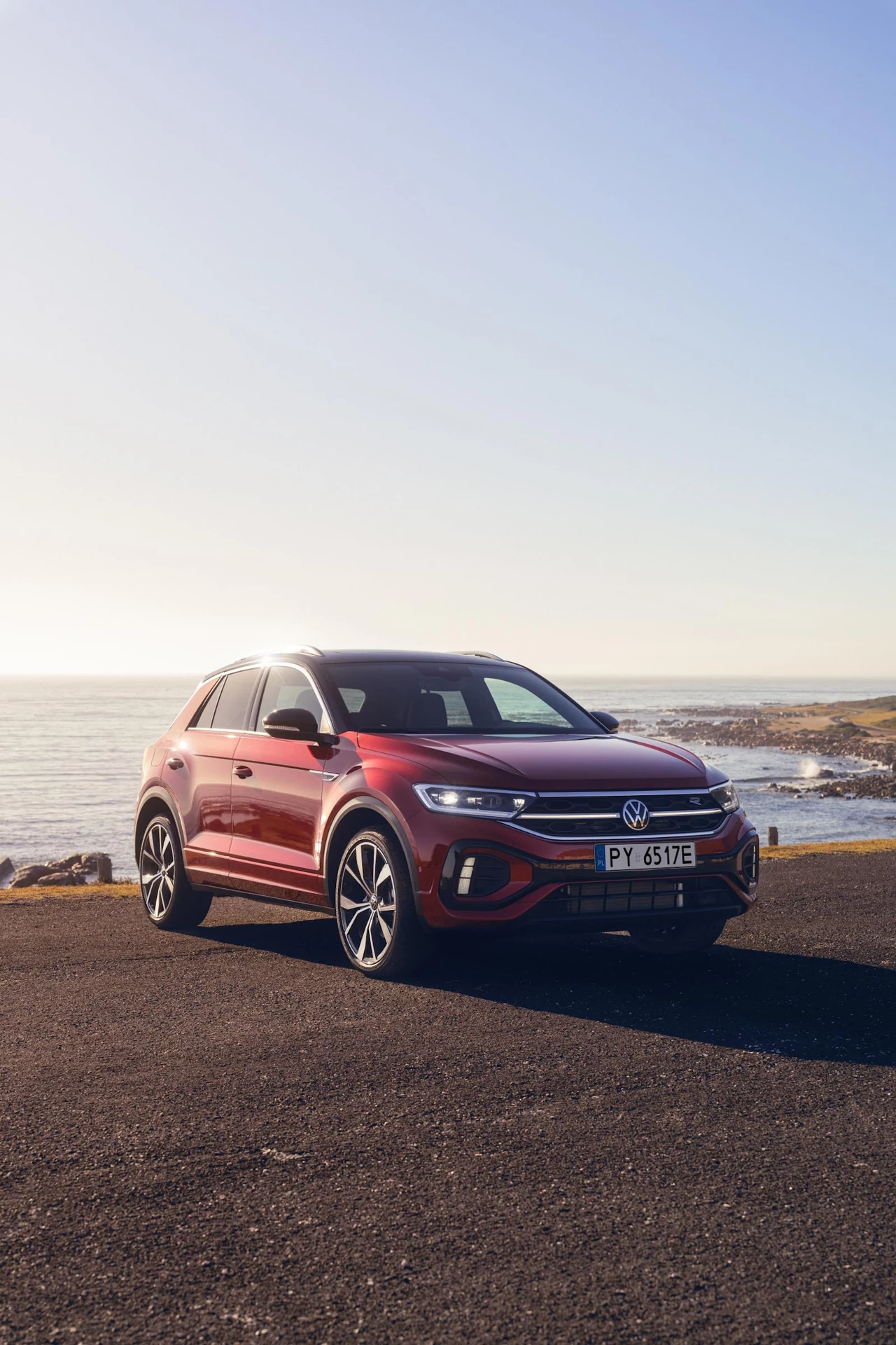 Volkswagen T-Roc został wyposażony w nowoczesną technologię, która nie tylko pomaga, ale i przewiduje Czerwony samochód marki Volkswagen zaparkowany na ciemnym asfalcie, w tle rozciąga się widok na morze i skalisty brzeg, niebo jest bezchmurne i jasne.