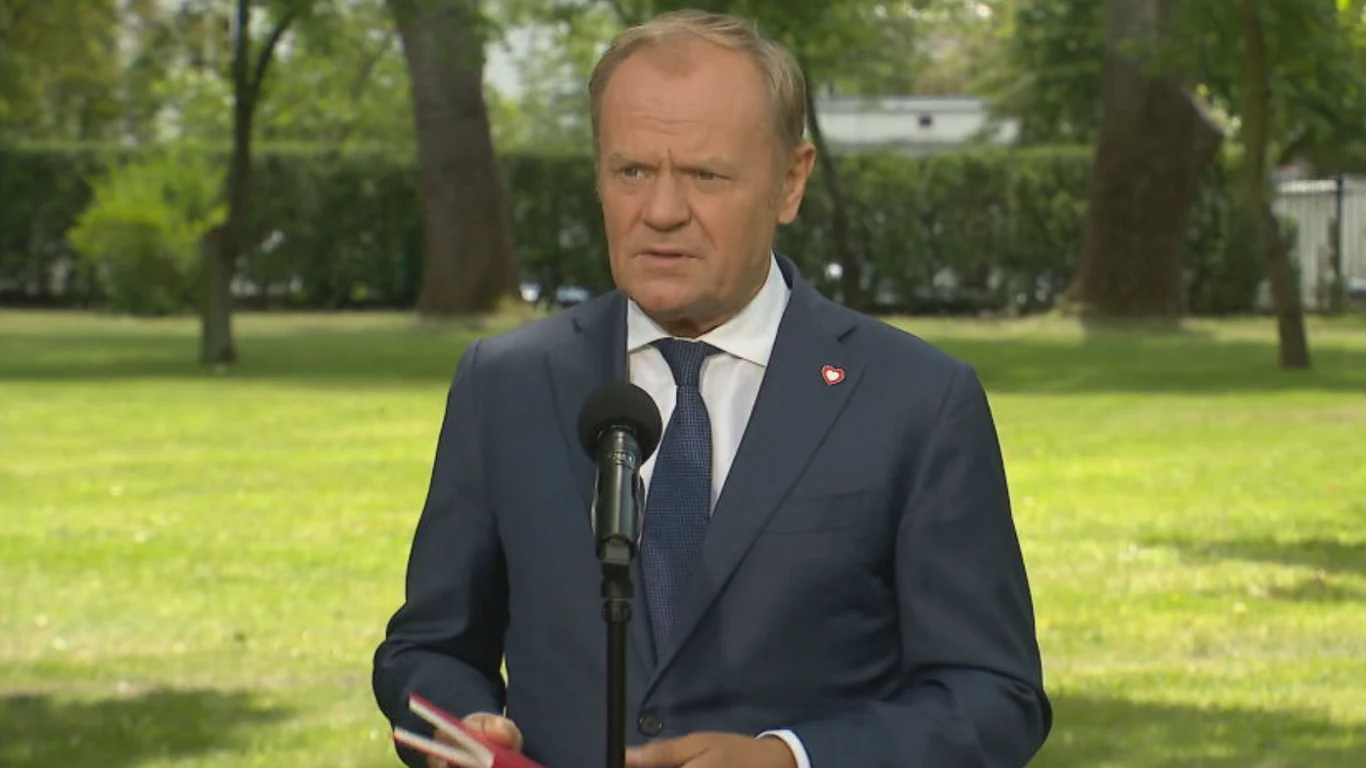 Donald Tusk Mężczyzna w garniturze stoi na tle zielonego parku, trzyma notatki i mówi do mikrofonu ustawionego przed nim, w tle widoczne drzewa i żywopłot.