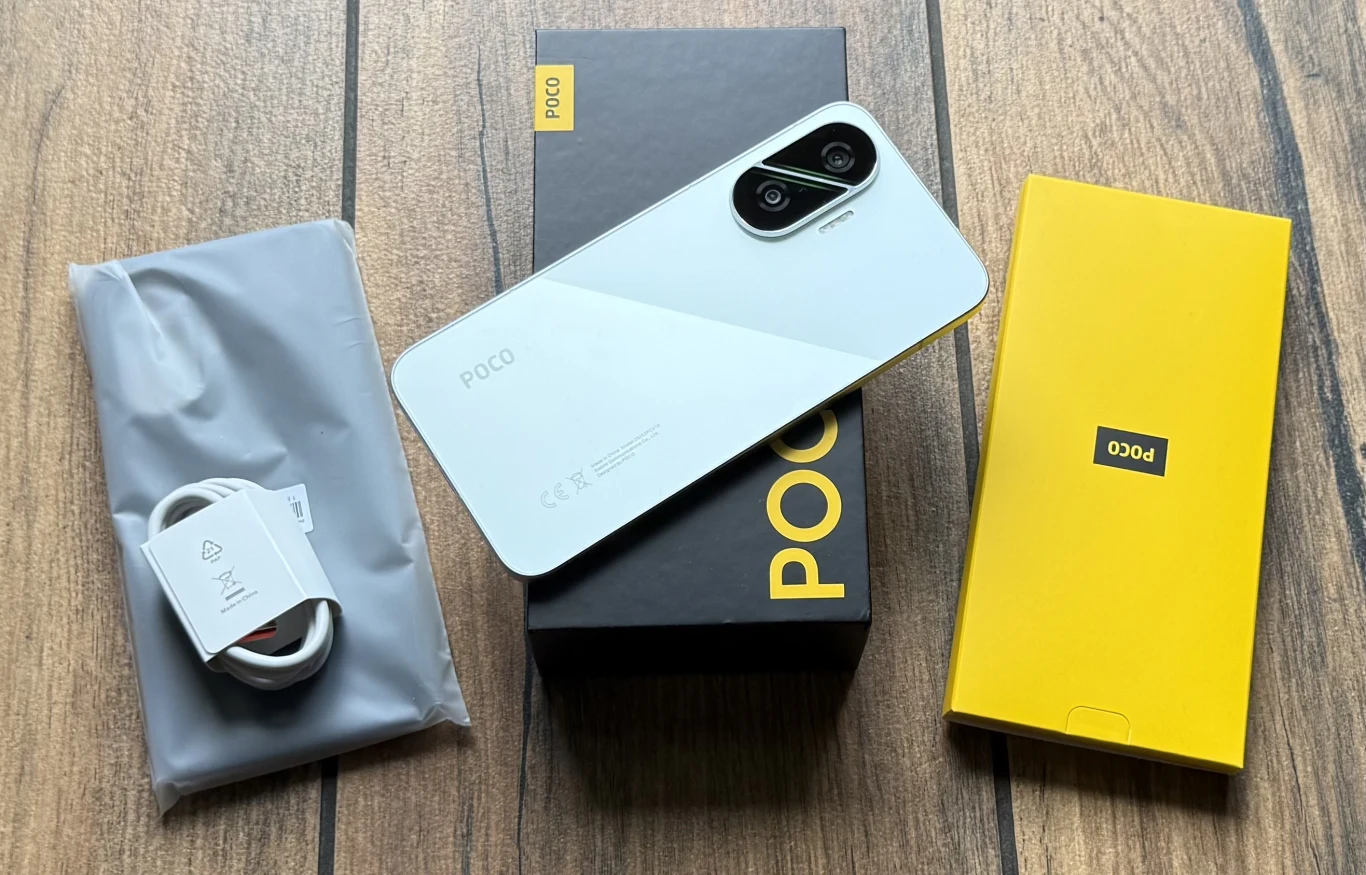 Nowoczesny smartfon POCO w białej obudowie, ułożony na czarnym pudełku z żółtym logo, obok rozpakowanego kabla USB z ładowarką oraz żółtego kartonika na drewnianej powierzchni.