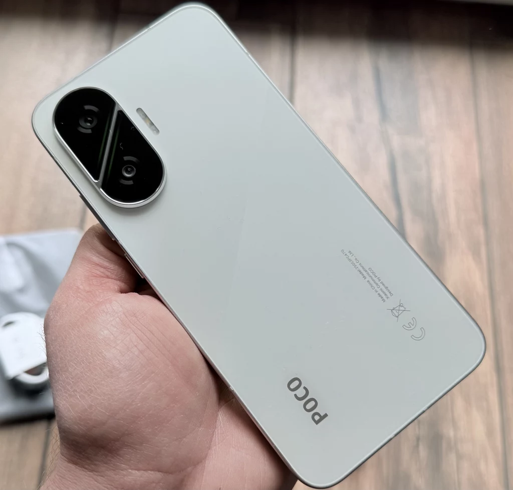 POCO F7 to smartfon, którego design przyciąga oko. Jasny smartfon marki POCO trzymany w dłoni, z widocznym podwójnym aparatem na tylnej obudowie, na tle drewnianej podłogi.