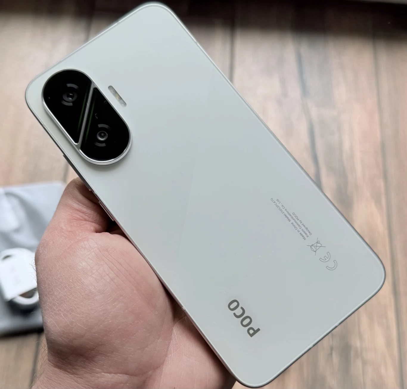 Jasny smartfon marki POCO trzymany w dłoni, z widocznym podwójnym aparatem na tylnej obudowie, na tle drewnianej podłogi.