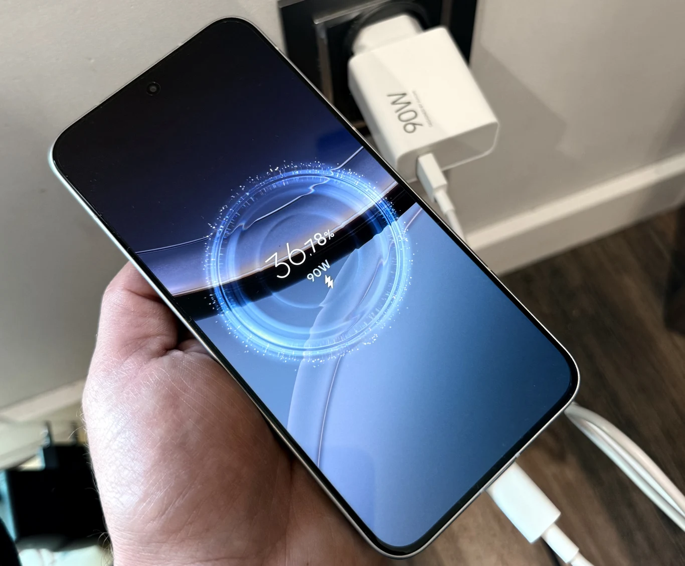 Nowoczesny smartfon trzymany w dłoni podczas ładowania, widoczny na wyświetlaczu dynamiczny wskaźnik naładowania baterii z informacją o szybkości ładowania, w tle biała ładowarka sieciowa podpięta do gniazdka.