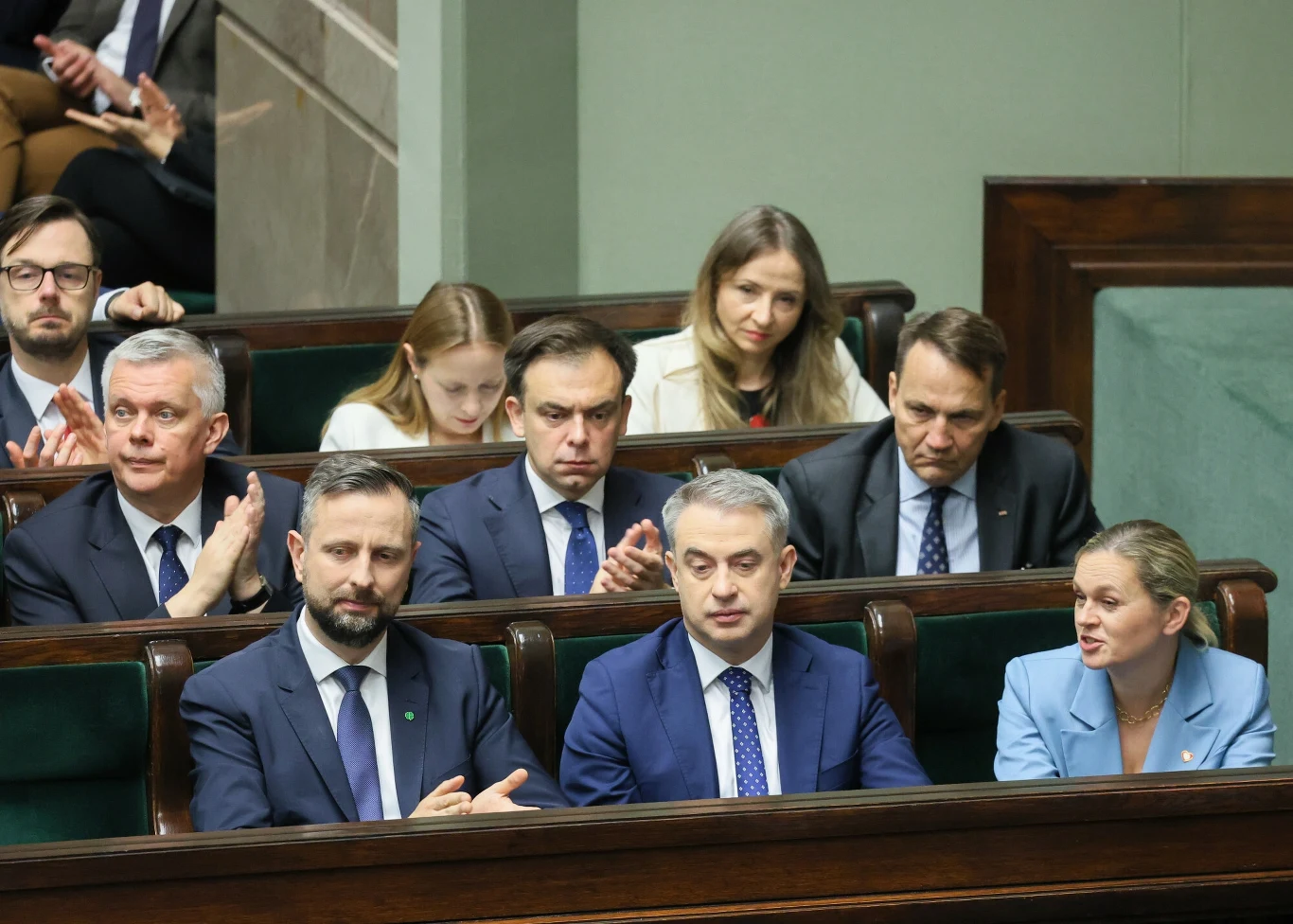 Rekonstrukcja rządu. Donald Tusk: Niech nikt nie śpi spokojnie Grupa polityków w garniturach siedzi w ławach parlamentarnych, część z nich rozmawia ze sobą, inni patrzą przed siebie lub biją brawo, dominują poważne miny i atmosfera skupienia.