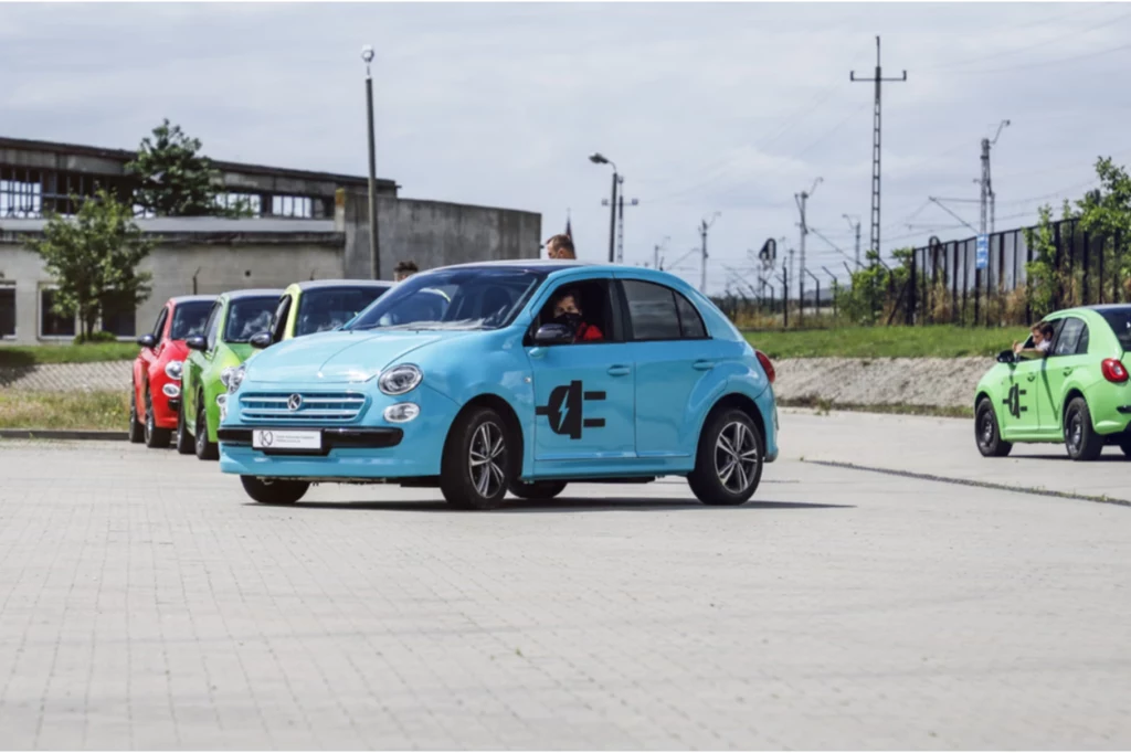 Elektryczne Vosco S106EV. Kolorowe samochody ustawione w rzędzie na otwartym placu z betonową nawierzchnią, na pierwszym planie niebieski samochód z oznaczeniami na drzwiach, w tle widać kolejne auta oraz zabudowania przemysłowe i elementy infrastruktury kolejowej.