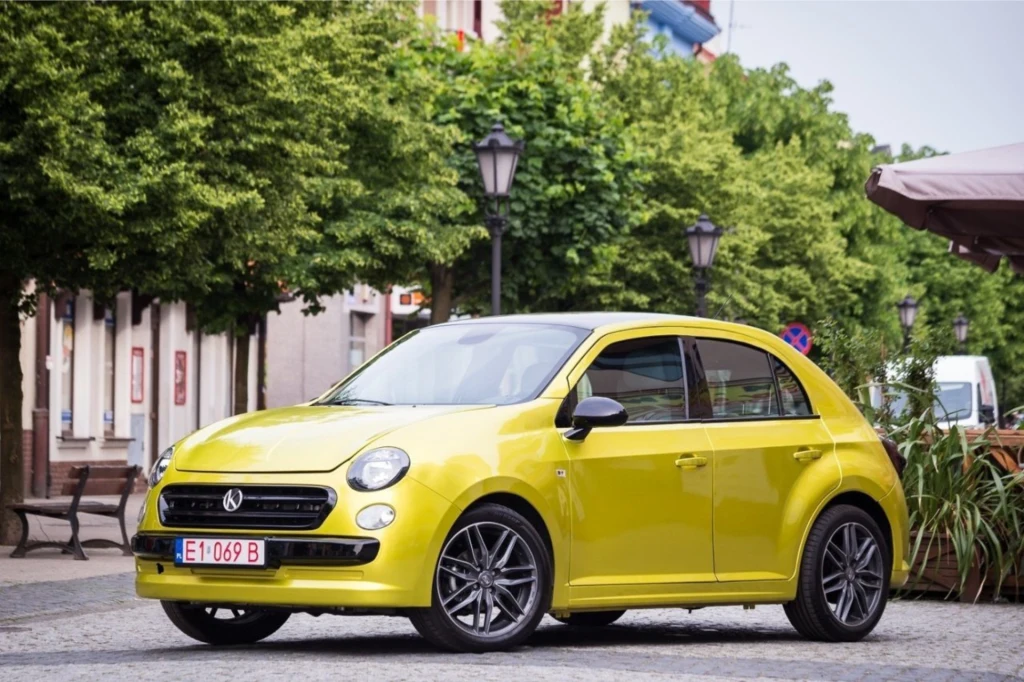 Jaskrawożółty samochód osobowy typu hatchback zaparkowany na brukowanej ulicy miejskiej, otoczony zielonymi drzewami oraz stylowymi latarniami, w tle widoczne fragmenty miejskiej zabudowy i sklepowe markizy.