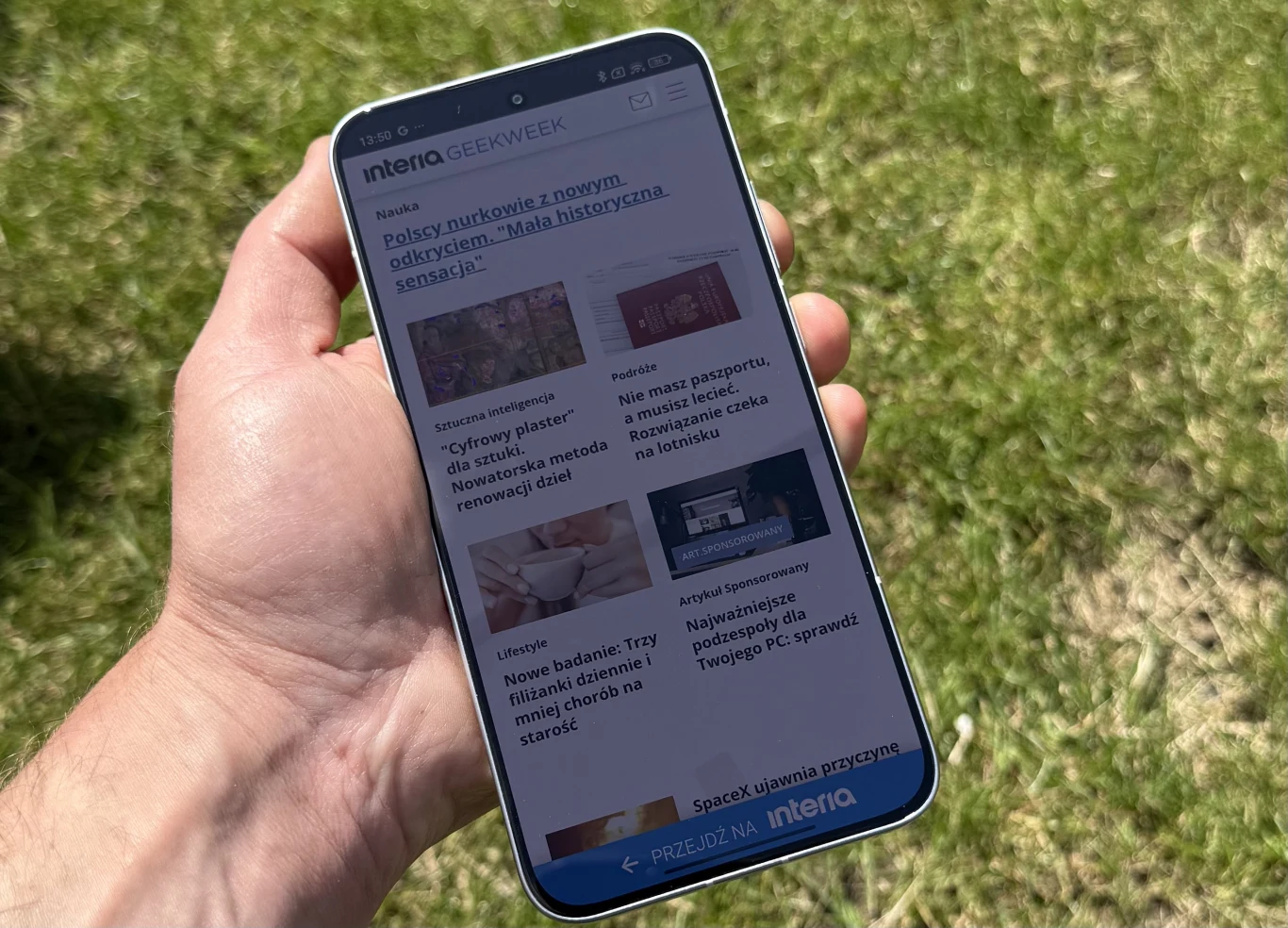 Ręka trzymająca smartfon z widoczną stroną internetową wyświetlaną na ekranie, w tle zielona trawa.