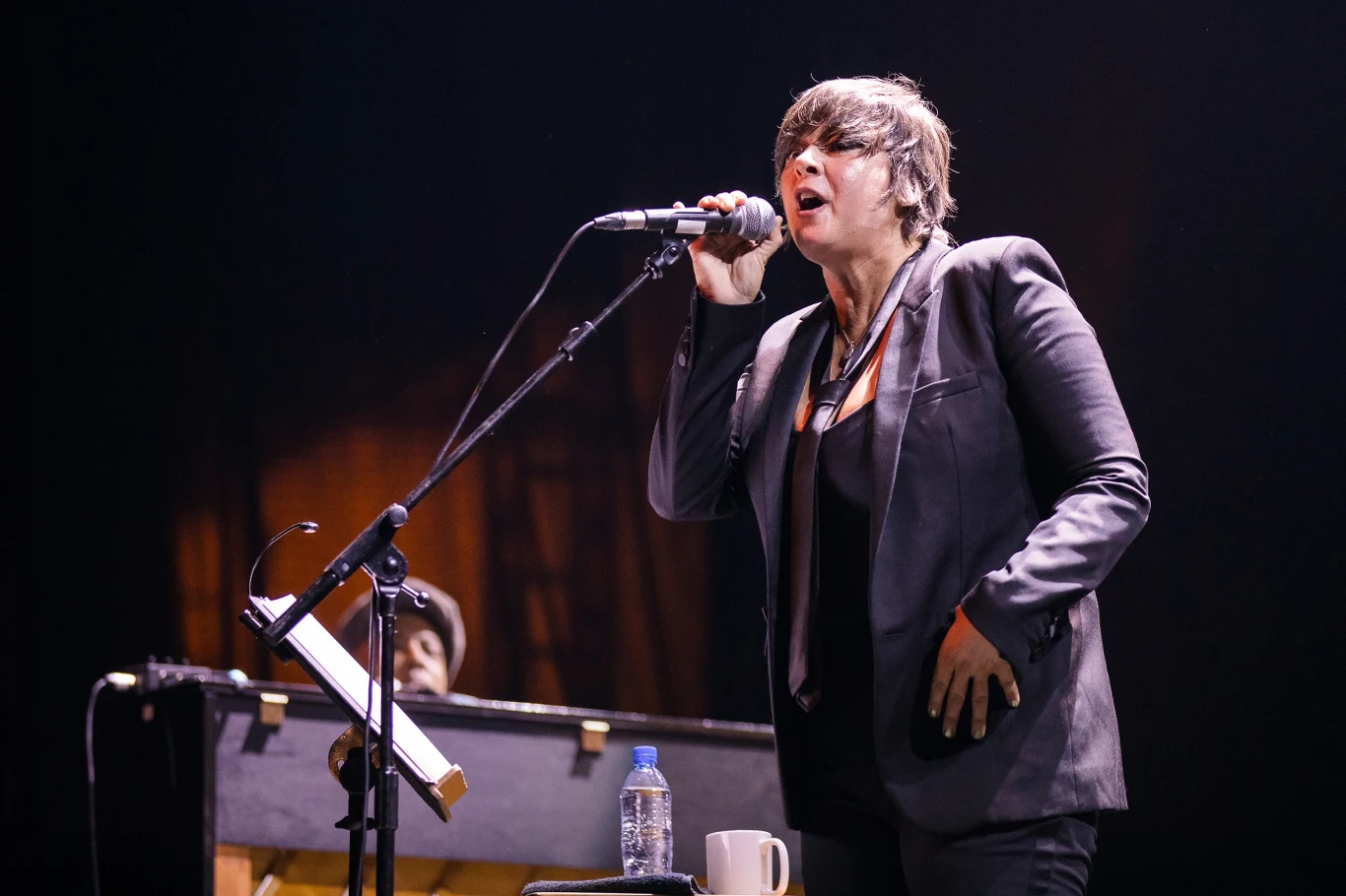 Cat Power wystąpiła na jednym koncercie w Polsce Kobieta w garniturze śpiewająca do mikrofonu na scenie, przy fortepianie, z butelką wody i kubkiem ustawionymi blisko nut.