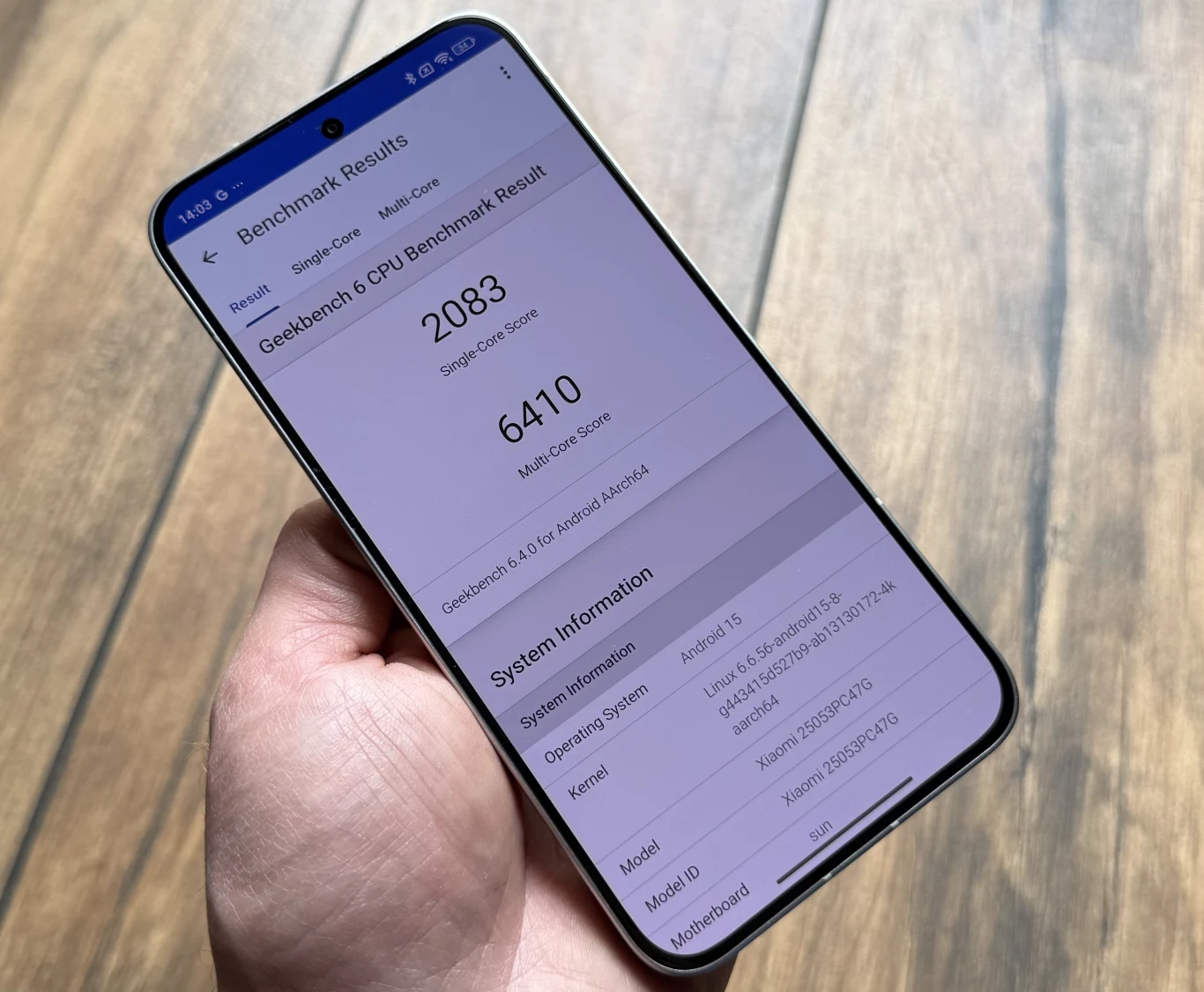Osoba trzyma w dłoni smartfon z wyświetlonym ekranem testu wydajności Geekbench 6, na którym widoczne są wyniki testu procesora i szczegółowe informacje o urządzeniu. Telefon znajduje się na tle drewnianej podłogi.