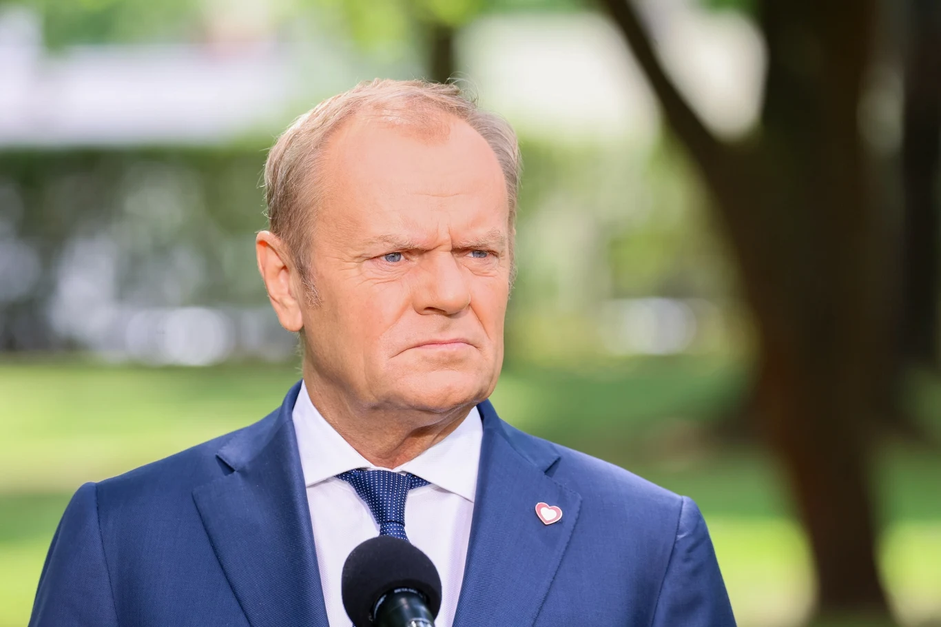 Donald Tusk Mężczyzna w garniturze i krawacie stoi na zewnątrz przed mikrofonem, z poważnym wyrazem twarzy, w tle rozmyte drzewa i zielona przestrzeń.