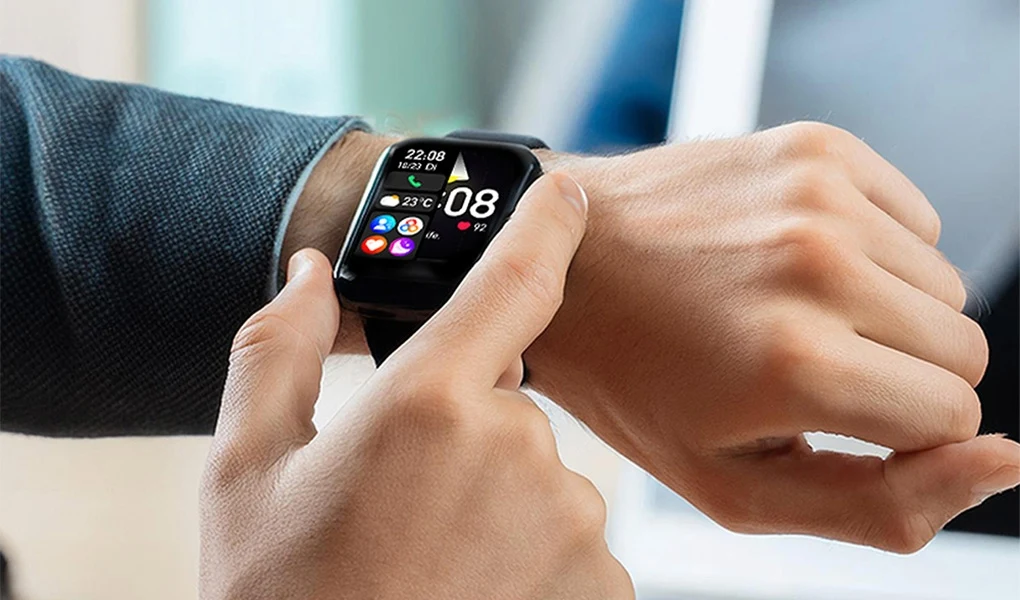 Smartwatch to dziś nie gadżet, a codzienne narzędzie Męska dłoń obsługująca nowoczesny smartwatch z wyświetlaczem dotykowym pokazującym godzinę, datę, pogodę oraz ikony aplikacji, w tle widoczny fragment ubrania i rozmyte elementy biura.