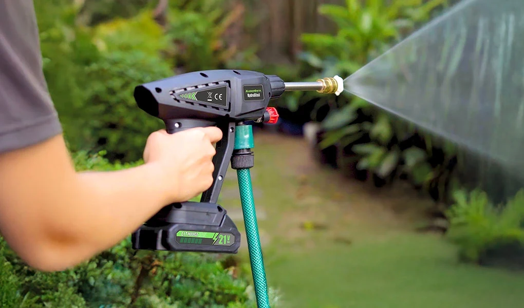 Hammersmith HydroBlast to precyzyjne narzędzie do zadań, które wymykają się tradycyjnym środkom czystości Ręka osoby trzymającej bezprzewodową myjkę ciśnieniową połączoną z ogrodowym wężem, urządzenie rozpyla wodę pod ciśnieniem na zielone rośliny w tle ogrodu.