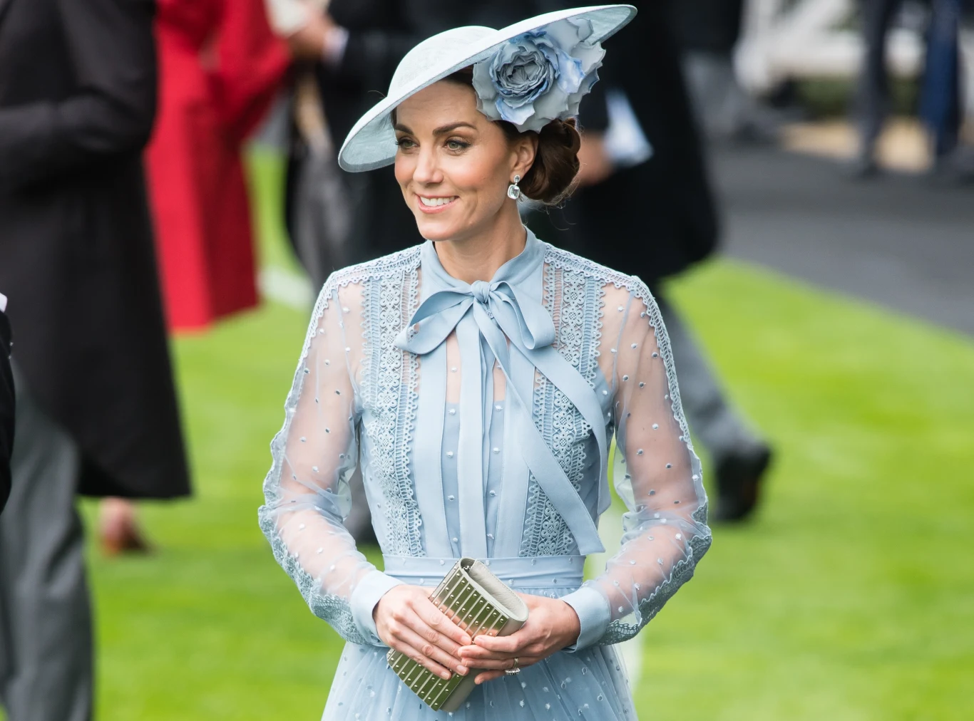 Baby blue króluje w szafie księżnej Kate Elegancko ubrana kobieta w błękitnej, ozdobionej koronkami sukni z długimi rękawami i szerokim kapeluszem z dekoracyjnym kwiatem, trzymająca w dłoni srebrną kopertówkę, spacerująca po zielonym trawniku wśród innych osób.