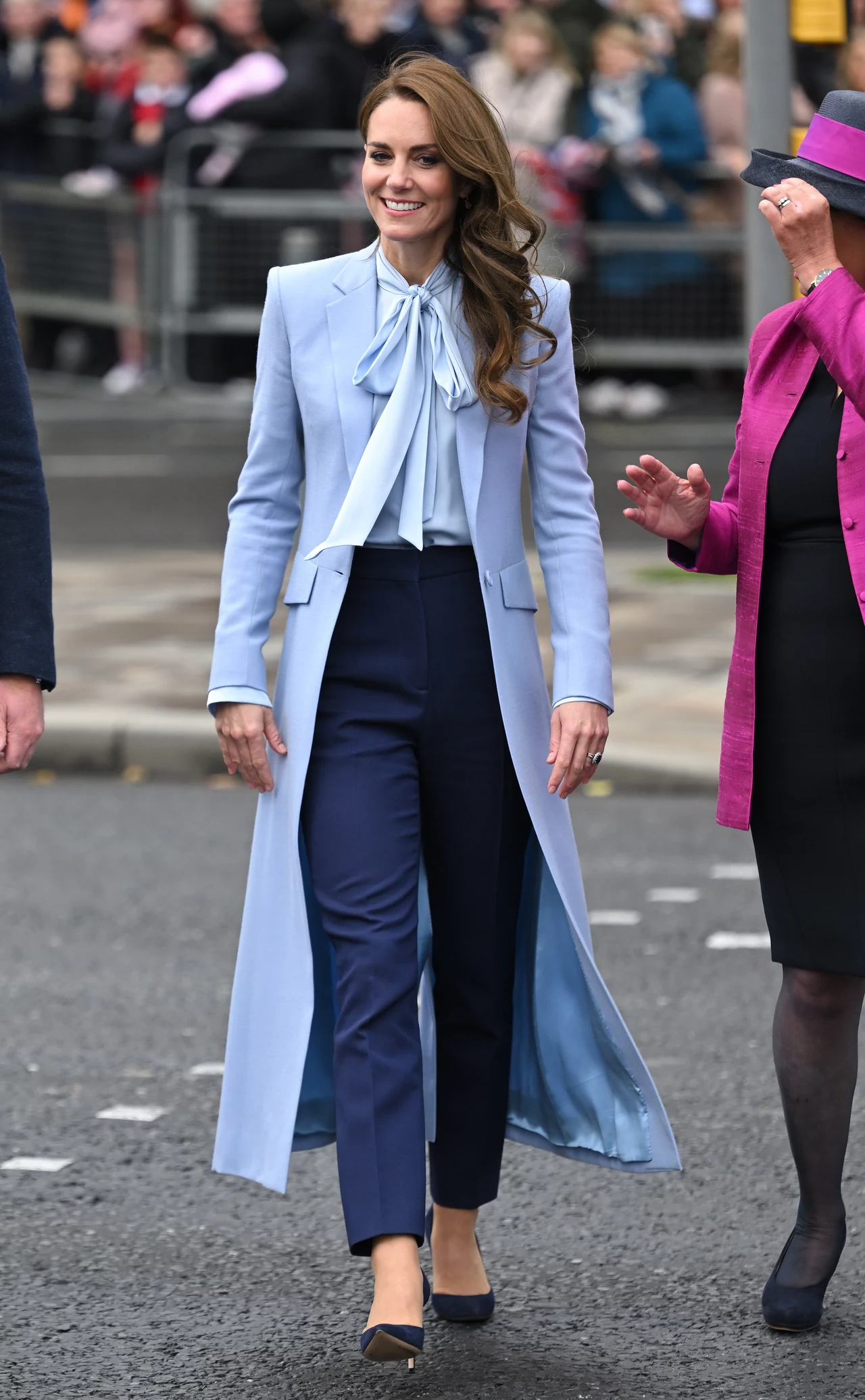 Błękitno-granatowa stylizacja z 2022 roku nadal jest w modowych trendach Kobieta w eleganckiej, jasnoniebieskiej marynarce z długim płaszczem, jasną bluzką z wiązaniem pod szyją i granatowych spodniach, uśmiecha się i spaceruje ulicą, w tle widoczni przechodnie oraz osoby zatrzymane barierkami.