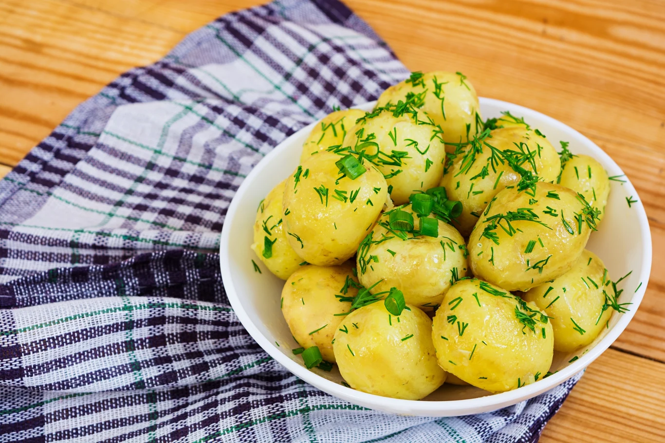 Miseczka świeżo ugotowanych młodych ziemniaków posypanych posiekanym koperkiem, ustawiona na kolorowej ściereczce kuchennej na drewnianym stole.