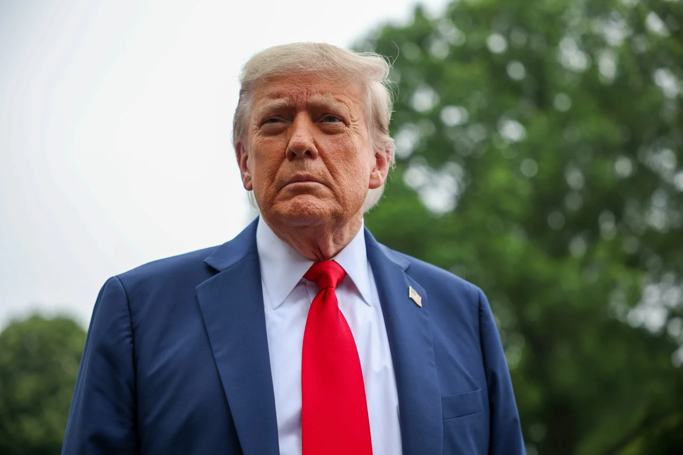 Donald Trump uważa, że Europa nie będzie mogła pomóc w zakończeniu konfliktu Izraela z Iranem Mężczyzna w niebieskim garniturze, białej koszuli i czerwonym krawacie, stoi na tle zielonych drzew spoglądając poważnie przed siebie.