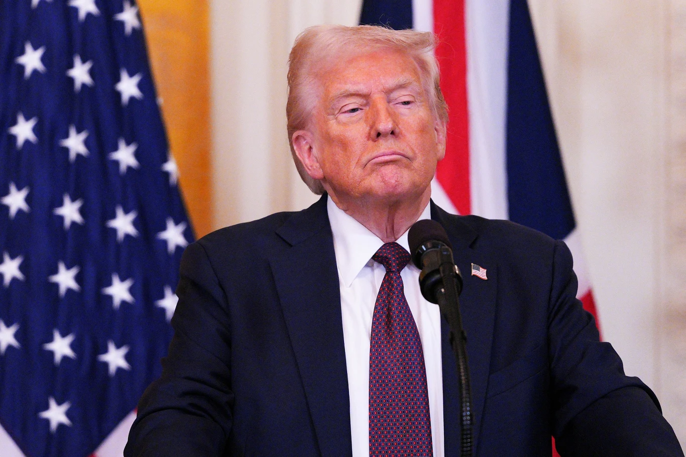 Donald Trump Mężczyzna w garniturze i czerwonym krawacie stoi przed mikrofonem na tle flag Stanów Zjednoczonych i Wielkiej Brytanii, jego twarz wyraża powagę i skupienie.