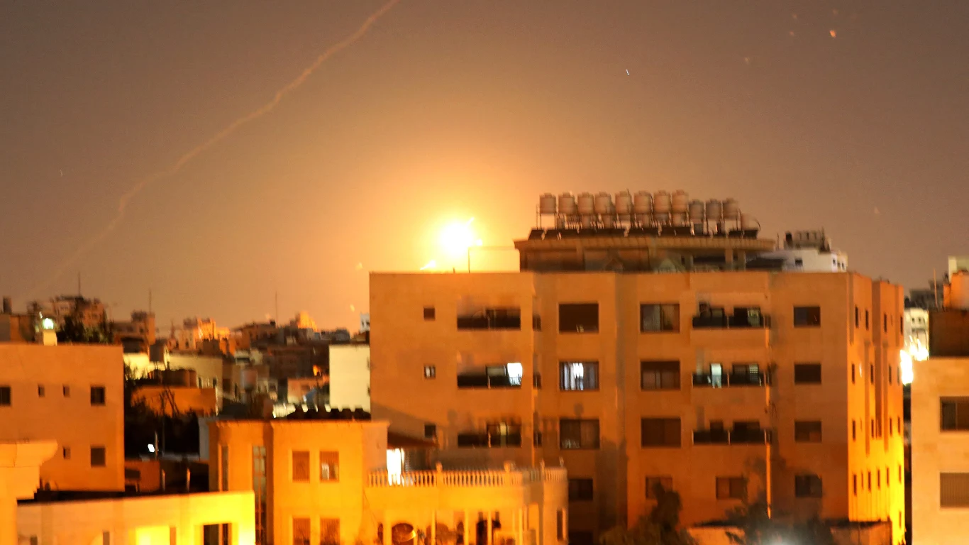 Izrael i Iran kontynuują ataki Wieczorna panorama miasta z jasnym rozbłyskiem na niebie za budynkami, sugerującym eksplozję lub wybuch w oddali, z widocznymi sylwetkami wielopiętrowych bloków mieszkalnych na pierwszym planie.