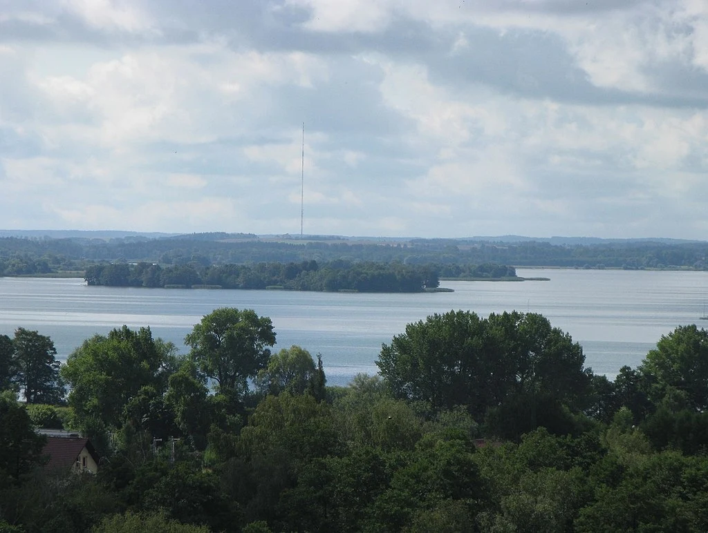 Jezioro otoczone gęstym lasem z wyspami pośrodku, w tle widoczny maszt nad taflą wody pod zachmurzonym niebem.