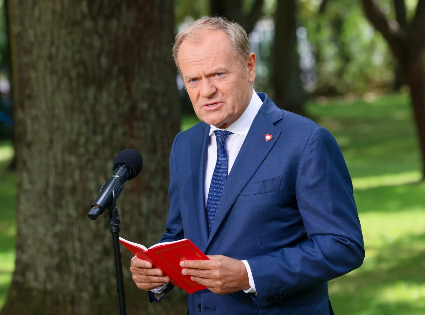 Donald Tusk zwrócił się z pytaniem do polityków PiS. Chodzi o wybory Mężczyzna w granatowym garniturze stojący na tle drzew, przemawiający do mikrofonu i trzymający w dłoniach czerwony notatnik.