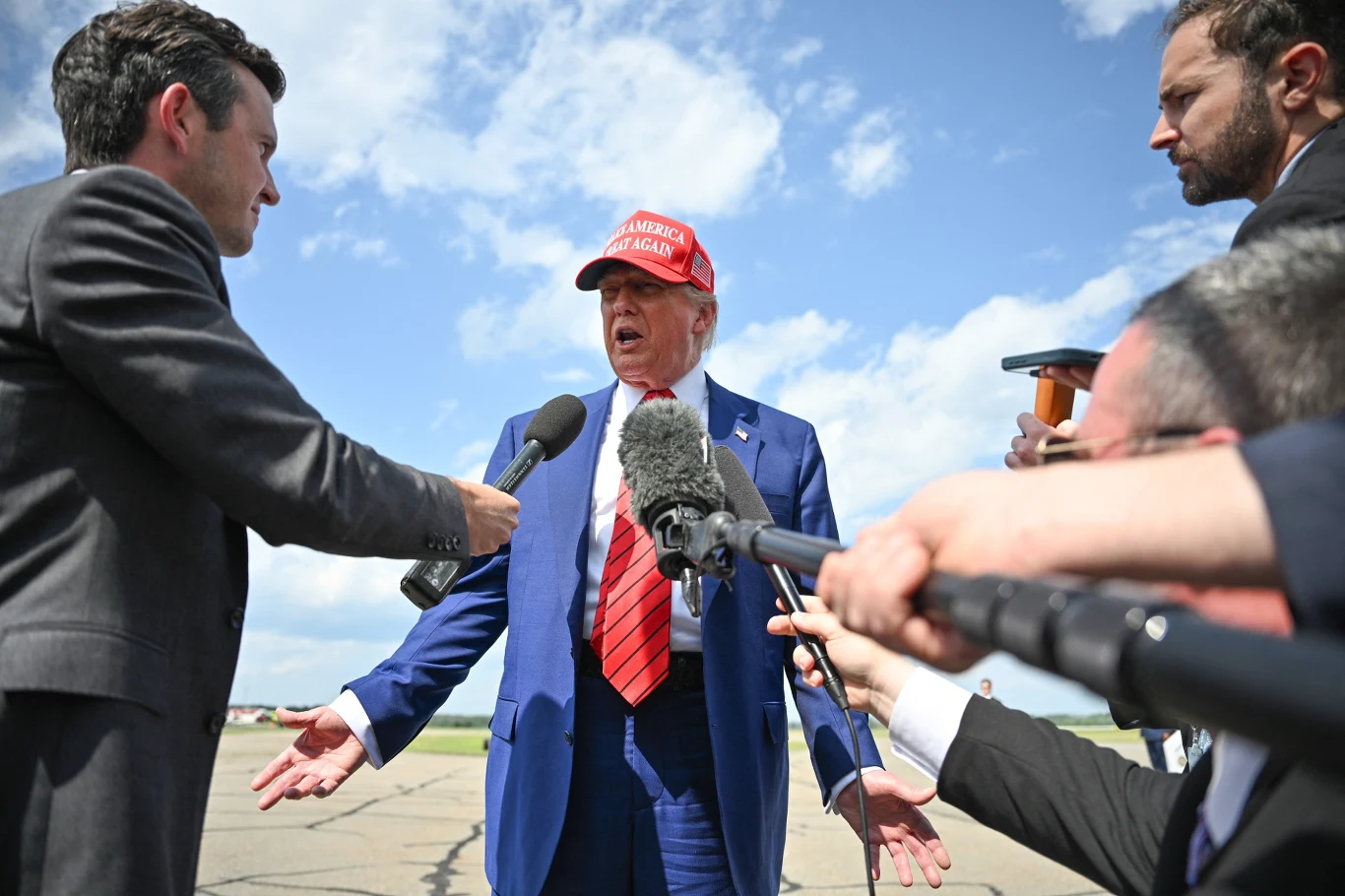 Mężczyzna w niebieskim garniturze i czerwonej czapce z napisem odpowiada na pytania dziennikarzy, wokół niego kilku reporterów trzyma mikrofony, wywiad prowadzony jest na tle nieba z chmurami.