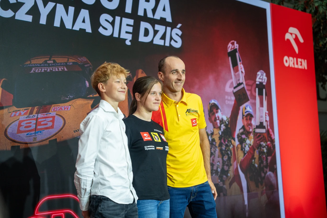 Podczas konferencji Robert Kubica poświęcił też nieco czasu młodym zawodnikom Akademii Orlen Teamu Trzy osoby pozujące razem na tle ścianki promocyjnej z logotypem Orlen oraz grafiką przedstawiającą zwycięzców wyścigu i samochód rajdowy Ferrari.