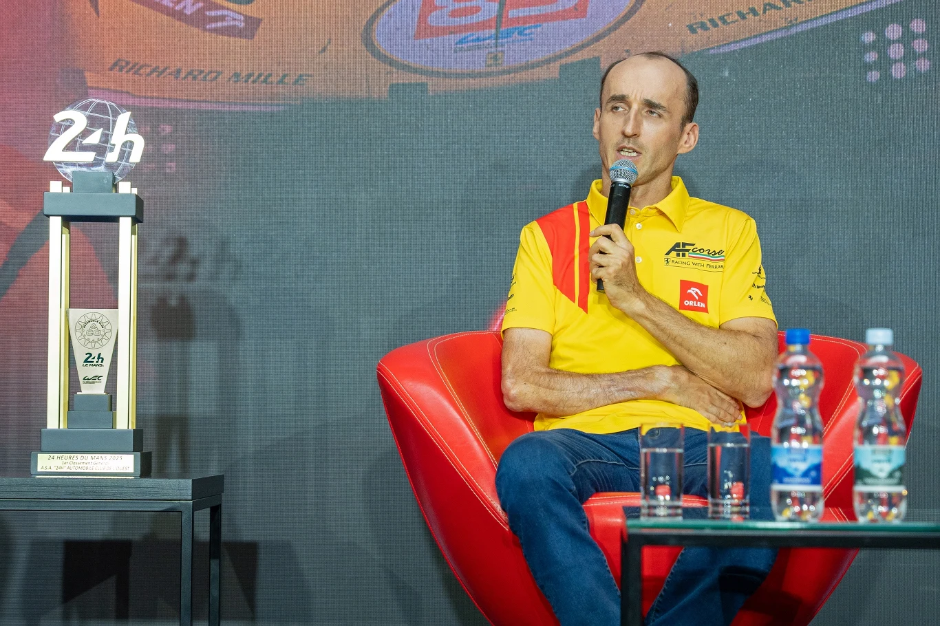 Robert Kubica kilka dni po zwycięstwie w Le Mans, pojawił się na konferencji w Warszawie Mężczyzna w żółtej koszulce z mikrofonem w ręku siedzi w czerwonym fotelu i udziela wywiadu, obok na stole stoi puchar. Na stole znajdują się także butelki wody i szklanki.