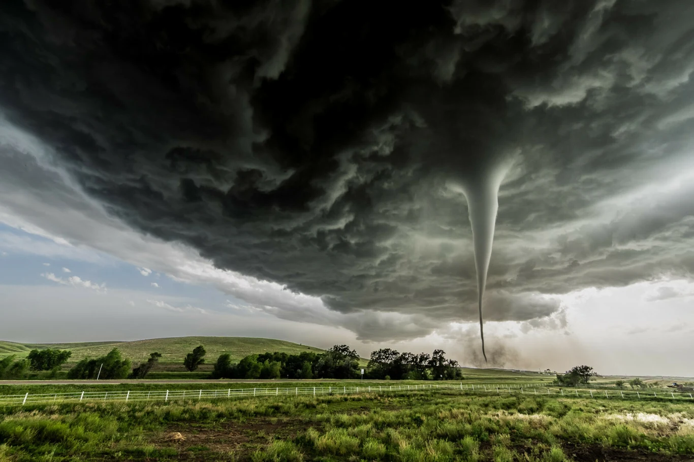 Na zdjęciu potężne tornado o prędkości wiatru przekraczającej 220 km/h, uchwycone przez brytyjskiego łowcę burz Davida Mayhew w pobliżu Dickens w stanie Nebraska, 16 czerwca 2025 r. Potężne tornado wiruje nad zielonym polem, pod ciemnymi, burzowymi chmurami; jasne światło rozświetla krajobraz na pierwszym planie, nadając dramatyczny efekt.