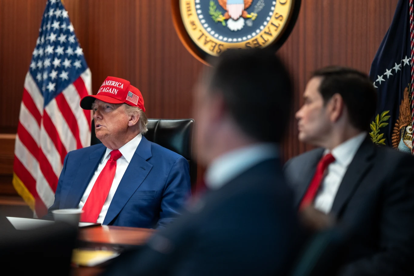 Donald Trump Mężczyzna w czerwonej czapce z napisem 'Make America Great Again' siedzi przy stole konferencyjnym w otoczeniu innych mężczyzn w garniturach, za nim widoczne amerykańskie flagi i pieczęć prezydencka.