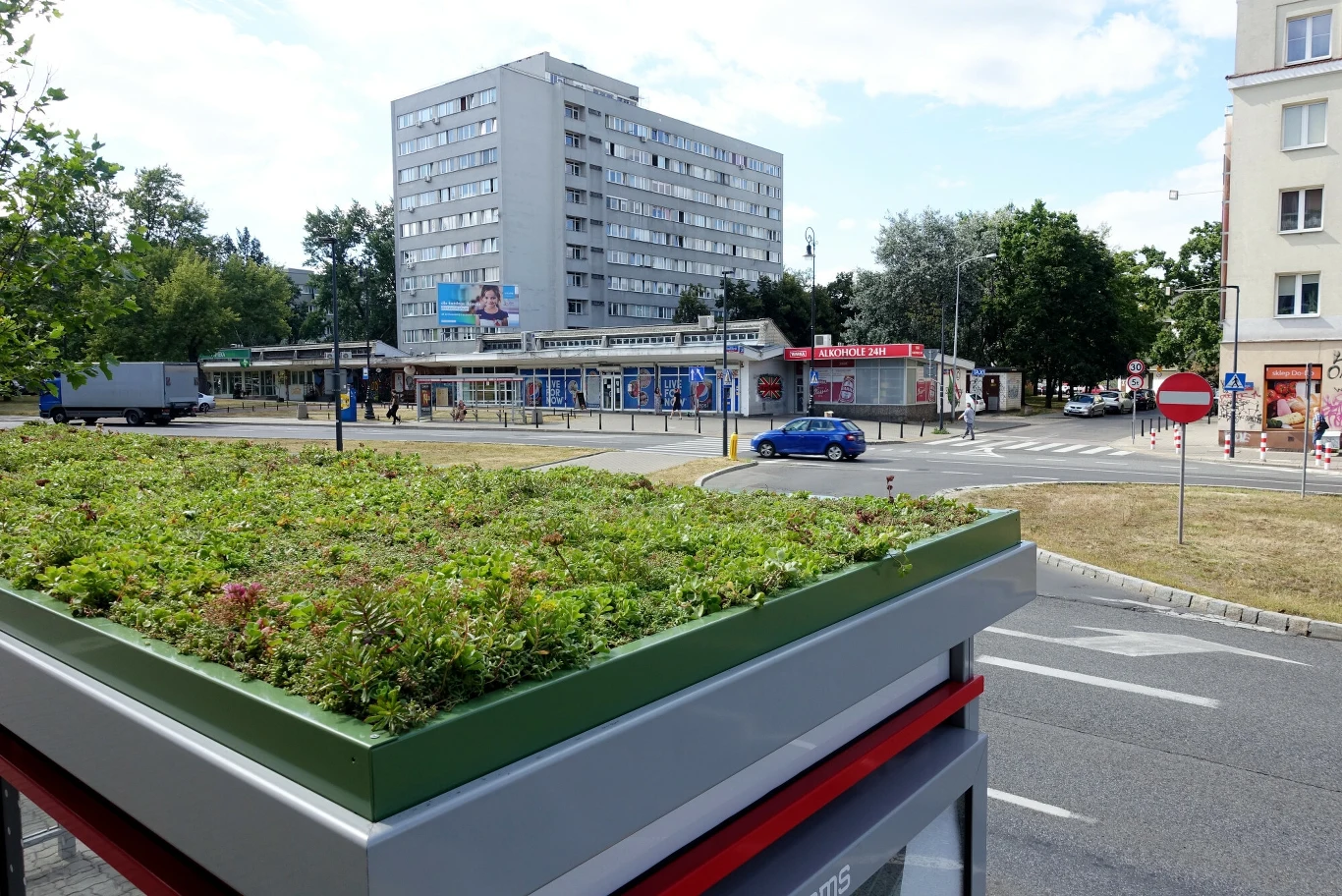 . To przyszłościowe rozwiązanie, choć jest kosztowne, to stanowi jedną z najciekawszych pozycji w zrównoważonym budownictwie przyszłości. Przystanek autobusowy z zielonym dachem na pierwszym planie, w tle ulica z samochodami, wysoki blok mieszkalny oraz niska zabudowa usługowa. Dookoła drzewa oraz fragment chodnika.