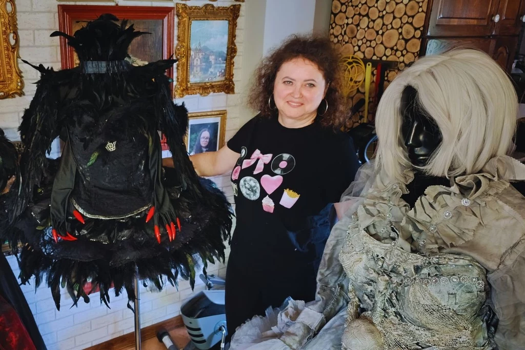 Elżbieta Tarłowska ze swoimi niezwykłymi sukniami. Czarną nazwała "Czarny łabędź" Kobieta z kręconymi włosami stoi w pomieszczeniu pomiędzy dwoma manekinami, na których znajdują się efektowne kostiumy – jeden czarny, ozdobiony piórami i czerwonymi detalami, drugi w jasnych, popielatych tonacjach, z bogatymi zdobieniami. W tle widocz...