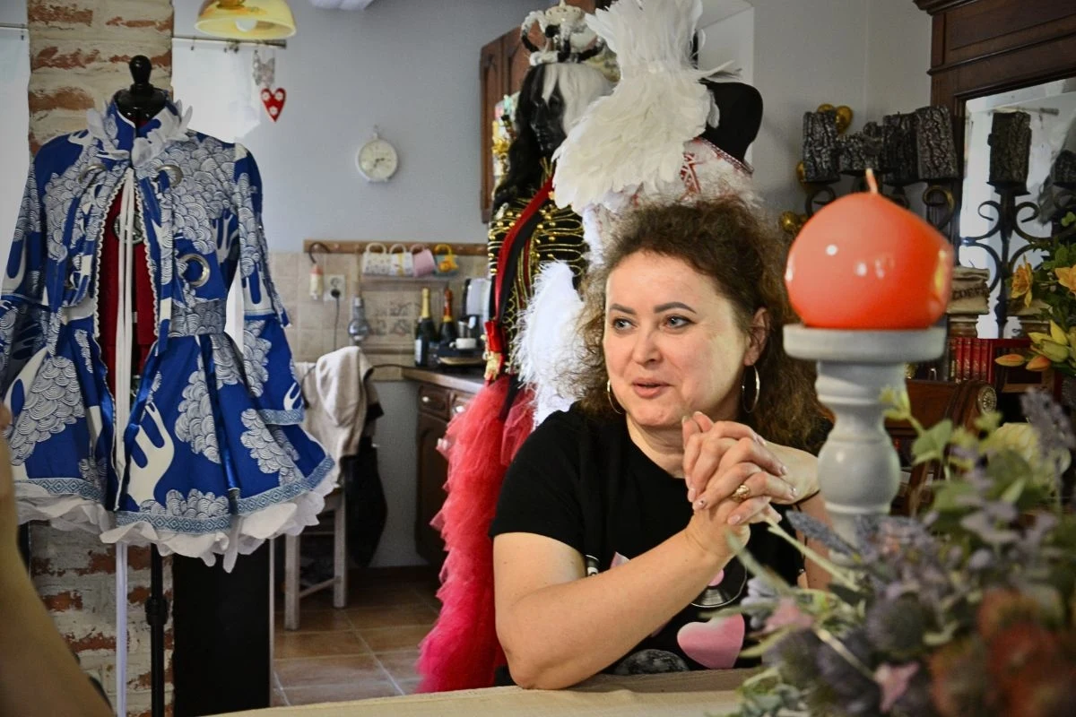 Kobieta siedząca przy stole w jasnym pomieszczeniu, w tle bogato zdobione kostiumy na manekinach oraz dekoracje charakterystyczne dla pracowni krawieckiej lub teatralnej.