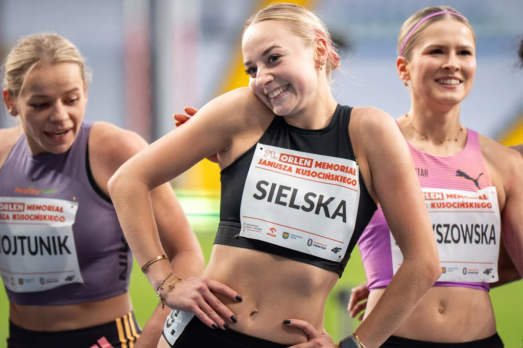 Alicja Sielska Trzy młode zawodniczki lekkoatletyczne w strojach sportowych z przypiętymi numerami, uśmiechnięte, stoją obok siebie na hali sportowej podczas wydarzenia sportowego